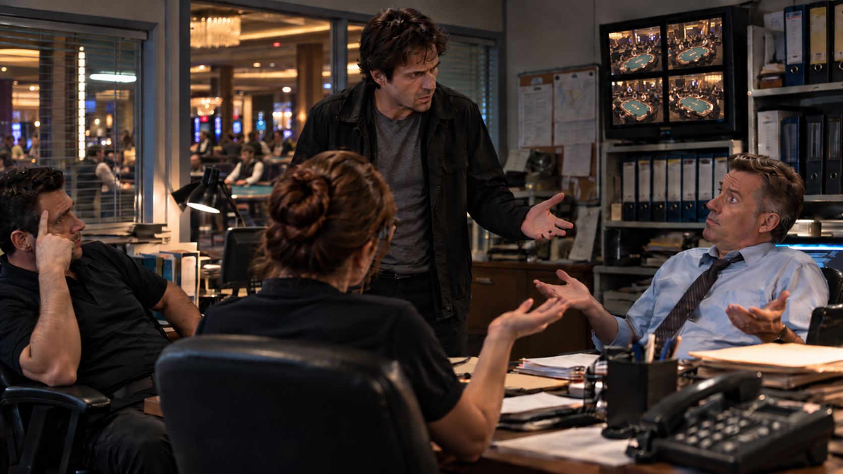 A confused casino surveillance technician questions his coworkers in the casino security office while surveillance monitors show poker tables in the gaming room behind them.